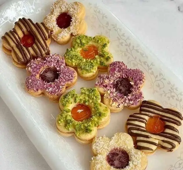 نمایی از شیرینی خانگی لذیذ و شیک، کوکیهای گلشکل رنگارنگ با مغز مربا و تزئینات پسته، نارگیل، شکلات و گل رز.
