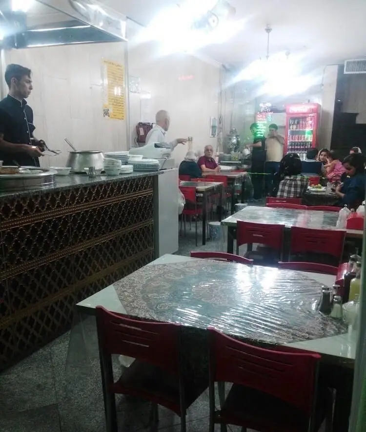 تصویر فضای داخلی طباخی بره ناقلا، شامل سرآشپز در حال آماده‌سازی و مشتریان در حال صرف غذای سنتی ایرانی.