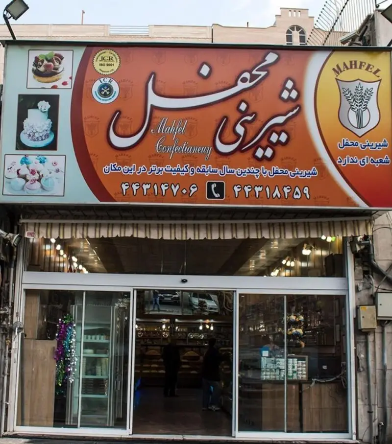 تصویر نمای بیرونی قنادی و شیرینی محفل با تابلوی بزرگ شامل تصاویر کیک، کاپ کیک، اطلاعات سابقه، شماره تماس و لوگوهای کیفیت.