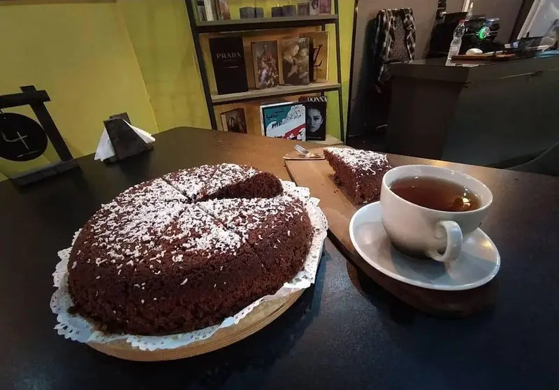 لذت چشیدن کیک شکلاتی بی‌نظیر کافه ژوانی با تزیین نارگیل و چای داغ در فضای دنج و کتابخانه‌ای این کافه.