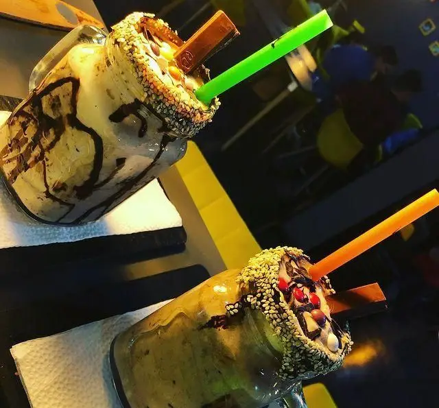 دو میلک شیک خلاقانه و خوشمزه کافه ژوانی با تاپینگ شکلات، کیت کت، دانه های کنجد و آبنبات رنگی در لیوان های شیشه‌ای، انتخابی عالی برای لحظات شیرین