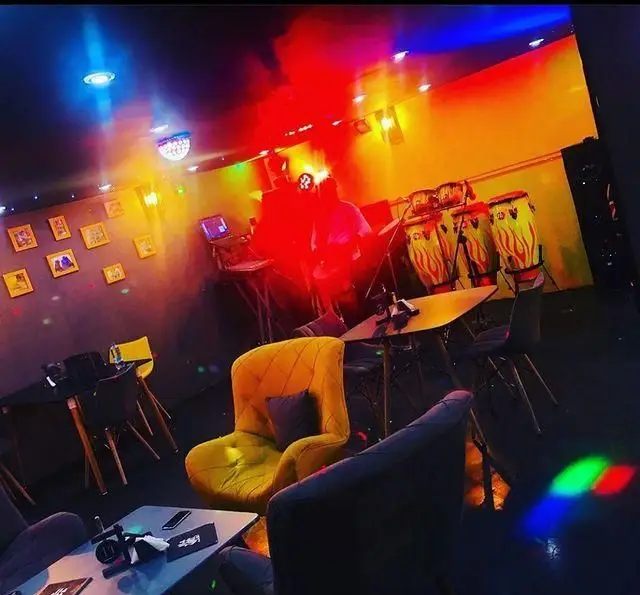 فضای داخلی کافه ژوانی با نورپردازی رنگارنگ و محیط شاد. شامل استیج موسیقی با درام و دی‌جی برای اجرای زنده.