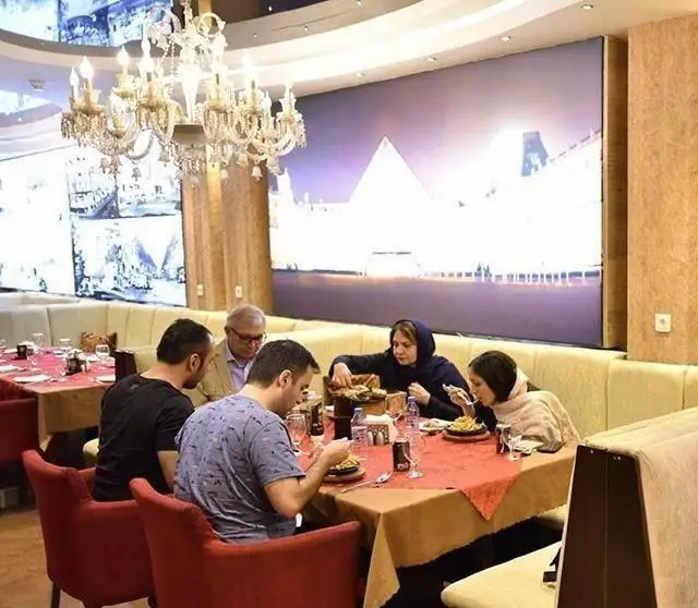 نمایی از دکوراسیون شیک رستوران طهران پاریس و مشتریان در حال صرف غذا در محیطی لوکس و پر نور با نمایشگرهای دیواری.