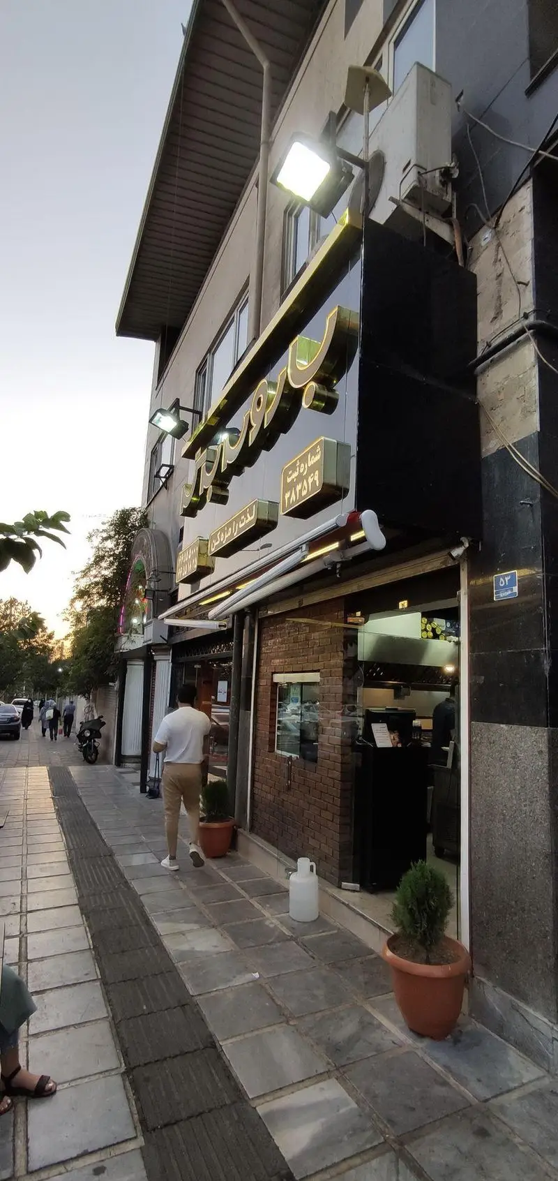 تصویری از نمای بیرونی رستوران باروس برگر با تابلوی بزرگ و حروف برجسته طلایی، شامل شماره ثبت و شعار "لذت را مزه کن"، در یک خیابان شهری ایران.