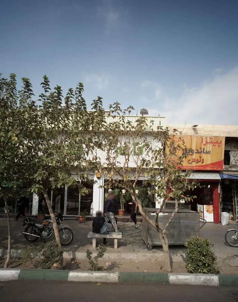 تصویری از نمای بیرونی کافه قنادی سعید در کنار خیابان، با درختان پوشیده از برگ، مشتریان نشسته و موتور سیکلت. روز آفتابی.