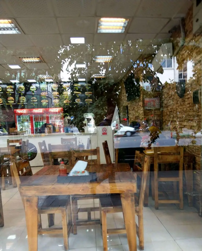 فضای داخلی فست فود ایران برگر: میز و صندلی چوبی، منوی برگر و نوشیدنی‌ها، با انعکاس خیابان و درختان از پشت ویترین.