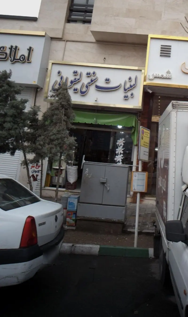 تصویری از نمای بیرونی مغازه لبنیات سنتی لیقوانی با تابلوی روشن، در کنار خیابان و خودروها، عرضه‌کننده محصولات لبنی محلی و تازه.