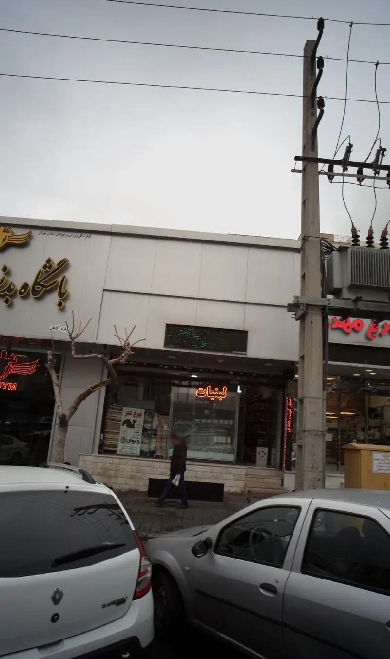 نمای خیابانی از فروشگاه لبنیات سنتی لیقوانی در تهران، شامل ویترین محصولات، تابلو مغازه، خودروهای پارک شده و عابران پیاده.