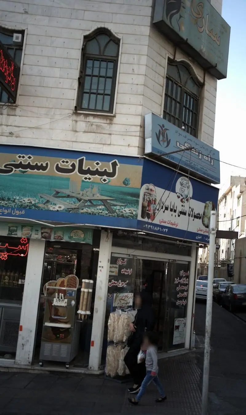 نمایی از فروشگاه لبنیات و محصولات سنتی باباسردار در یک خیابان شلوغ، با دستگاه بستنی ساز و عرضه شیرینی‌های محلی.