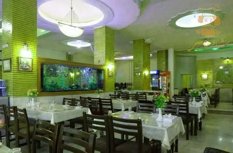 سالن غذاخوری رستوران شاطر عباس با دکوراسیون سبز، میز و صندلی چوبی و آکواریوم بزرگ دیواری، محیطی دلنشین برای صرف غذا.