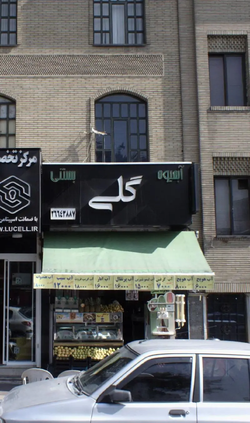 عکس از نمای بیرونی آبمیوه و بستنی گلی با تابلوی روشن، ویترین شیشه‌ای میوه‌های تازه و دستگاه آبمیوه گیری، و منوی قیمت محصولات.