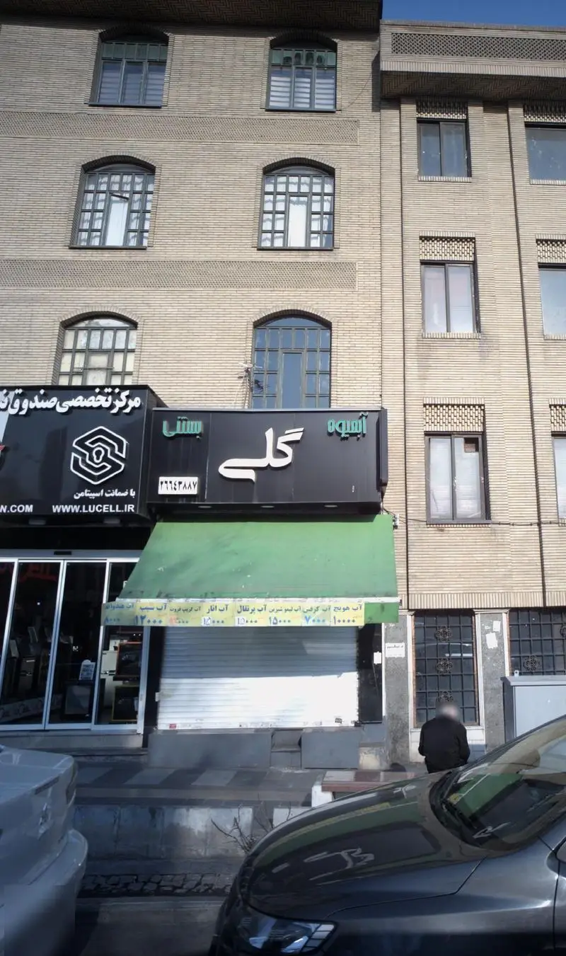 عکس از نمای بیرونی مغازه آبمیوه و بستنی گلی، شامل تابلو بزرگ فروشگاه، منوی نوشیدنی‌ها و بستنی‌ها با قیمت، و ساختمان آجری.