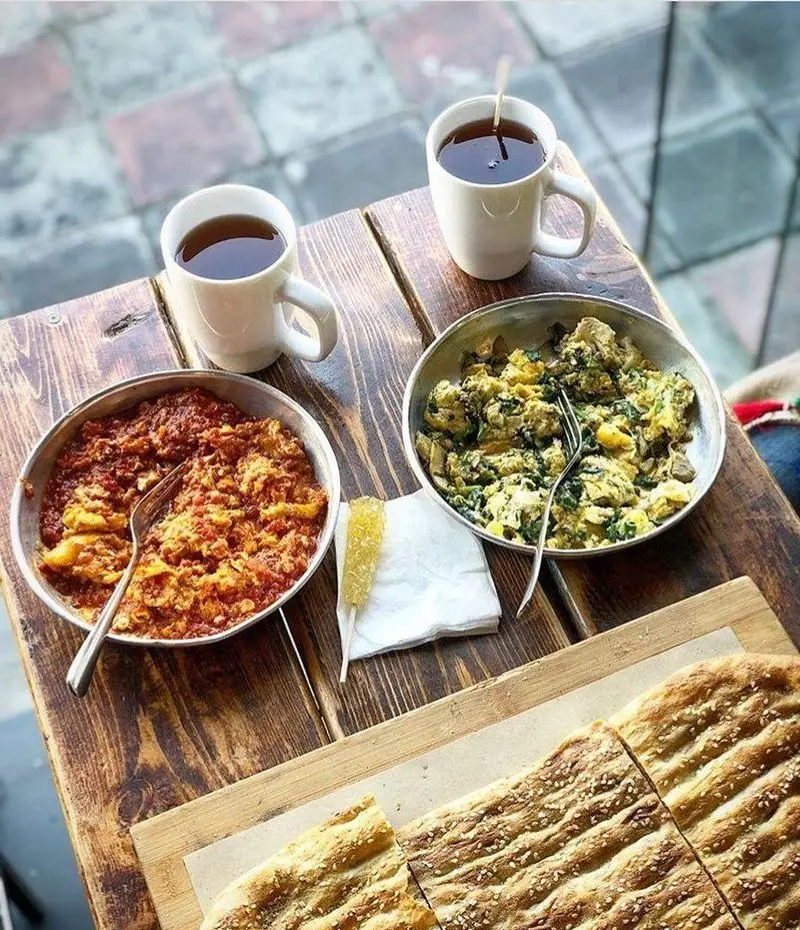 میز صبحانه کامل و خوشمزه در رستوران برادران شامل نیمرو، املت سبزیجات، نان کنجدی تازه و چای داغ ایرانی.