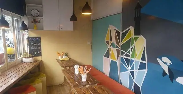 تصویری از فضای داخلی فست فود رگا، شامل میزهای چوبی دنج، دکوراسیون رنگارنگ مدرن، و تابلوی منوی پیتزا و پاستای ایتالیایی.