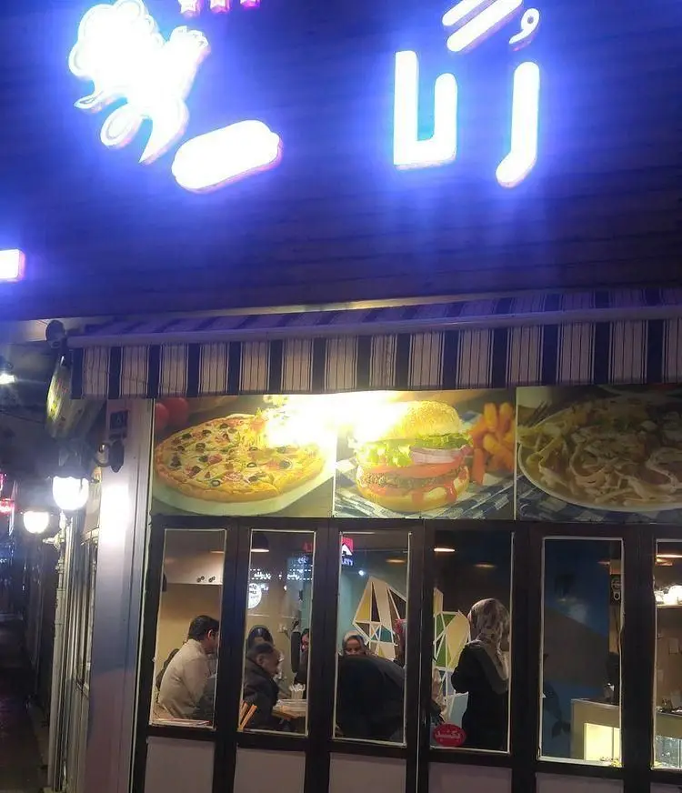 نمایی از فست فود رگا در شب با تابلوی نئونی درخشان، تصاویر پیتزا، همبرگر و پاستا در ویترین، و مشتریان داخل رستوران.