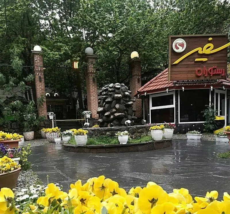فضای دلنشین رستوران سفیر، شامل آبنمای سنگی، باغچه پر از گل‌های زرد، ورودی با ستون‌های آجری و درختان سرسبز.