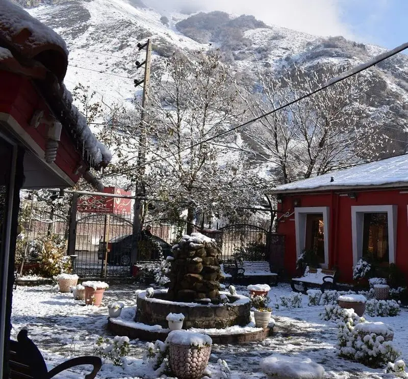 رستوران سفیر در زمستان: حیاط دلپذیر پوشیده از برف، فواره سنگی مرکزی، گلدان‌های برفی و کوهستان سفیدپوش در دوردست.