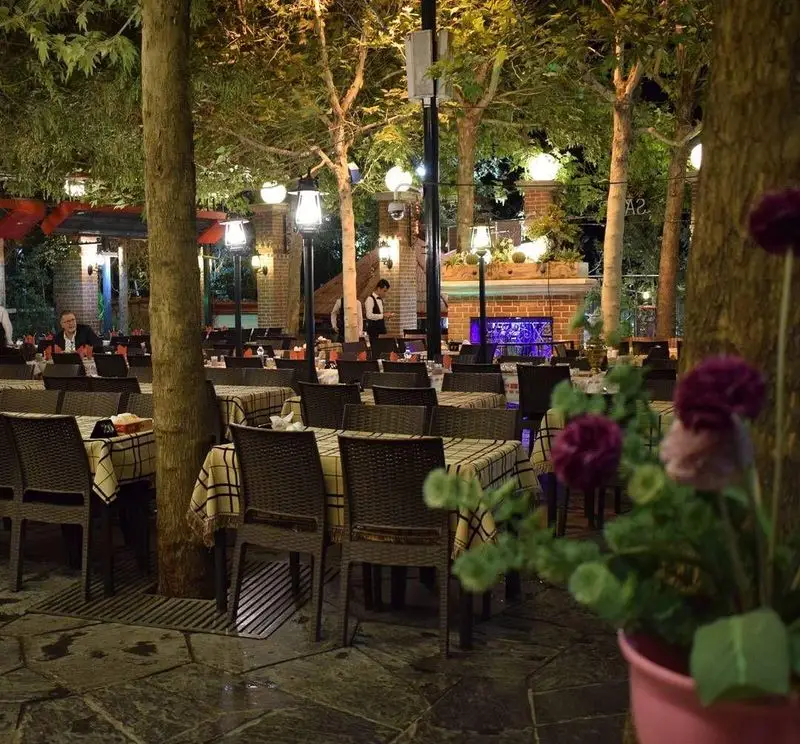 نمای شب از فضای باز و دلنشین رستوران سفیر، با میز و صندلی‌های چیده شده در میان درختان و نورپردازی زیبا. محیطی آرام و جذاب برای تجربه غذایی لذت‌بخش.
