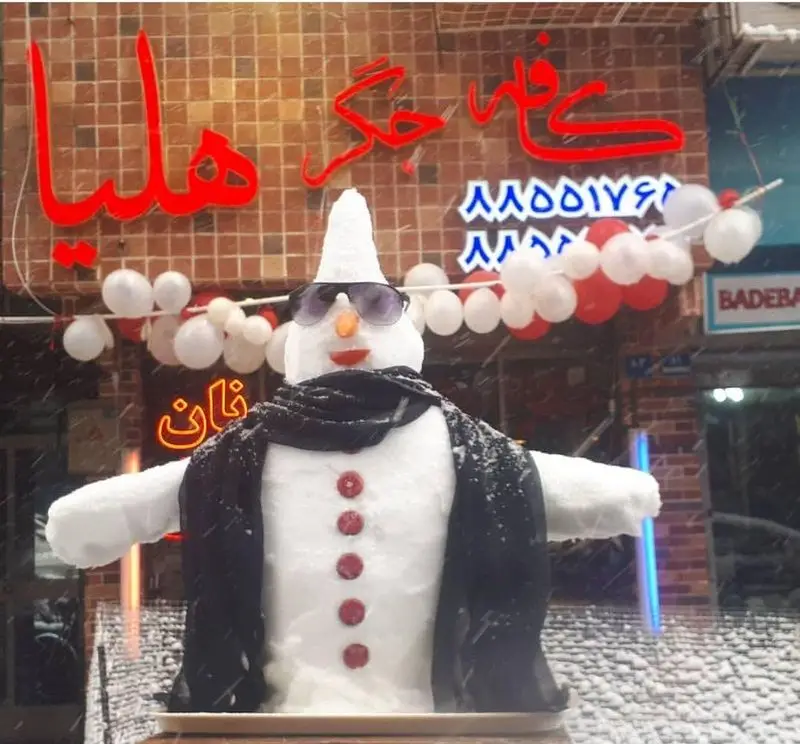 آدم برفی جذاب با عینک آفتابی و شال مشکی، در روز برفی مقابل کافه جگر هلیا. تزیینات بادکنکی قرمز و سفید و نام کافه هلیا با فونت بزرگ در پس زمینه.