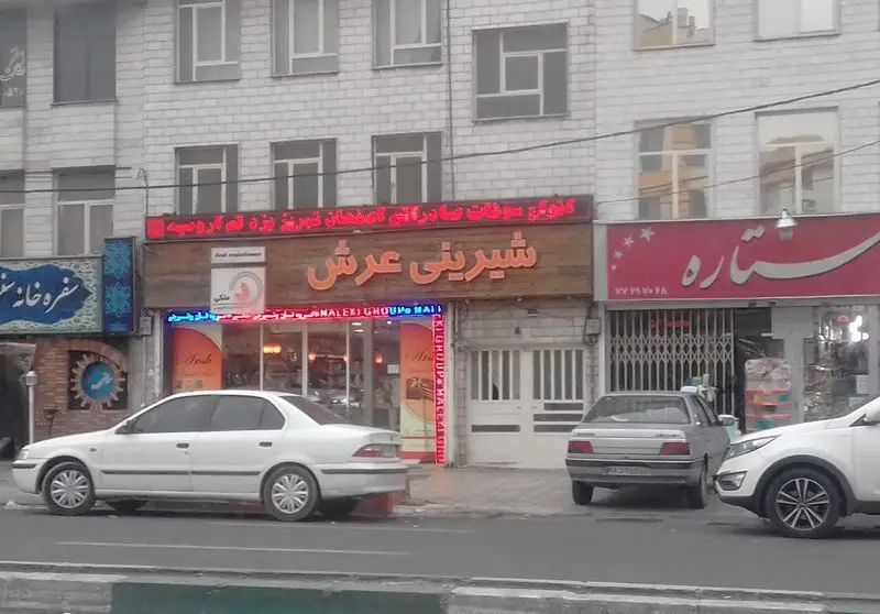 نمای بیرونی شیرینی عرش تهرانپارس، فروشگاه قنادی با تابلو چوبی بزرگ، در کنار سفره خانه و مغازه ستاره در خیابان.