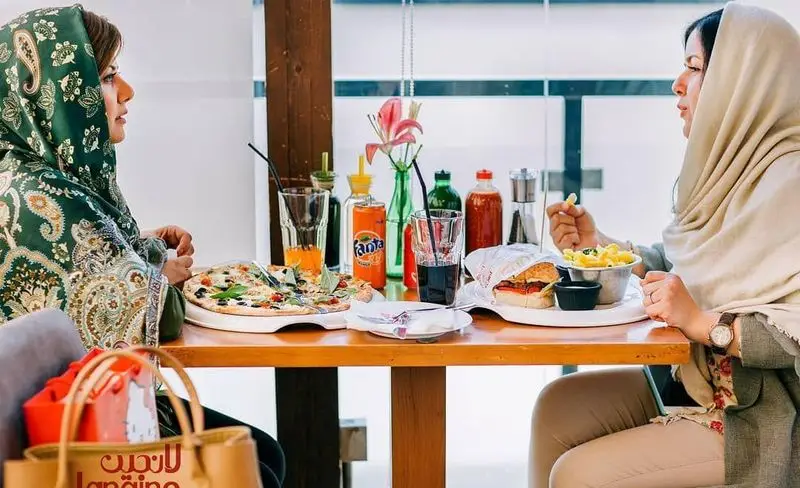 دو خانم مشتری در رستوران و کافه لانجین مشغول صرف غذاهای خوشمزه شامل پیتزا و برگر، در محیطی شاد و دوستانه هستند.