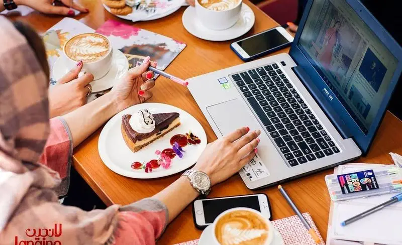 شخصی در کافه و رستوران لانجین با لپ‌تاپ کار می‌کند، در کنار قهوه لاته، کیک شکلاتی و دسرهای خوشمزه در محیطی دنج و آرام.