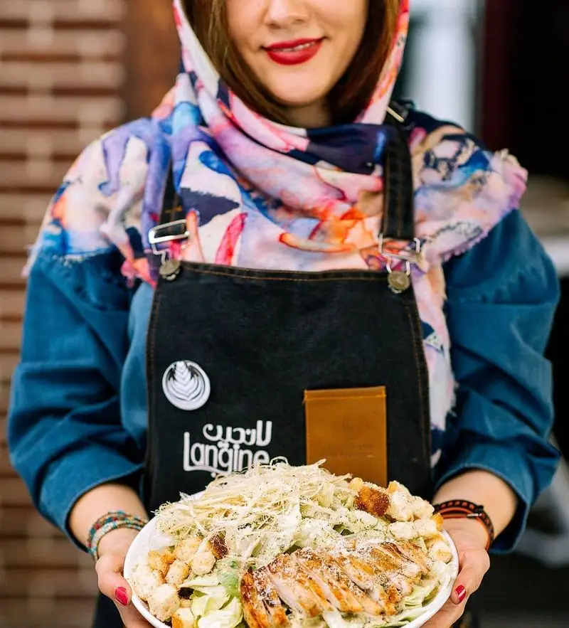 زنی در کافه لانجین با پیش‌بند لانجین، سالاد سزار با مرغ گریل خوشمزه و تازه را سرو می‌کند. تجربه غذای باکیفیت در رستوران لانجین.