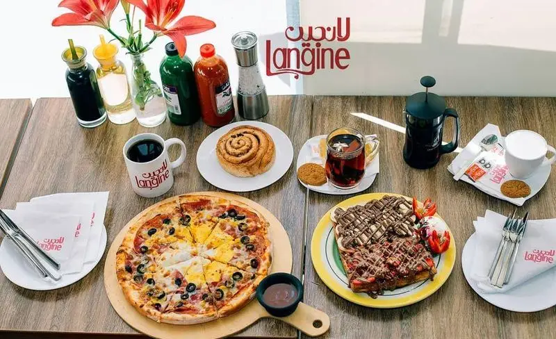 میز پر از غذاهای لذیذ و متنوع در رستوران و کافه لانجین شامل پیتزا، وافل شکلاتی توت فرنگی، رول دارچین و قهوه، چای و نوشیدنی‌های گرم.