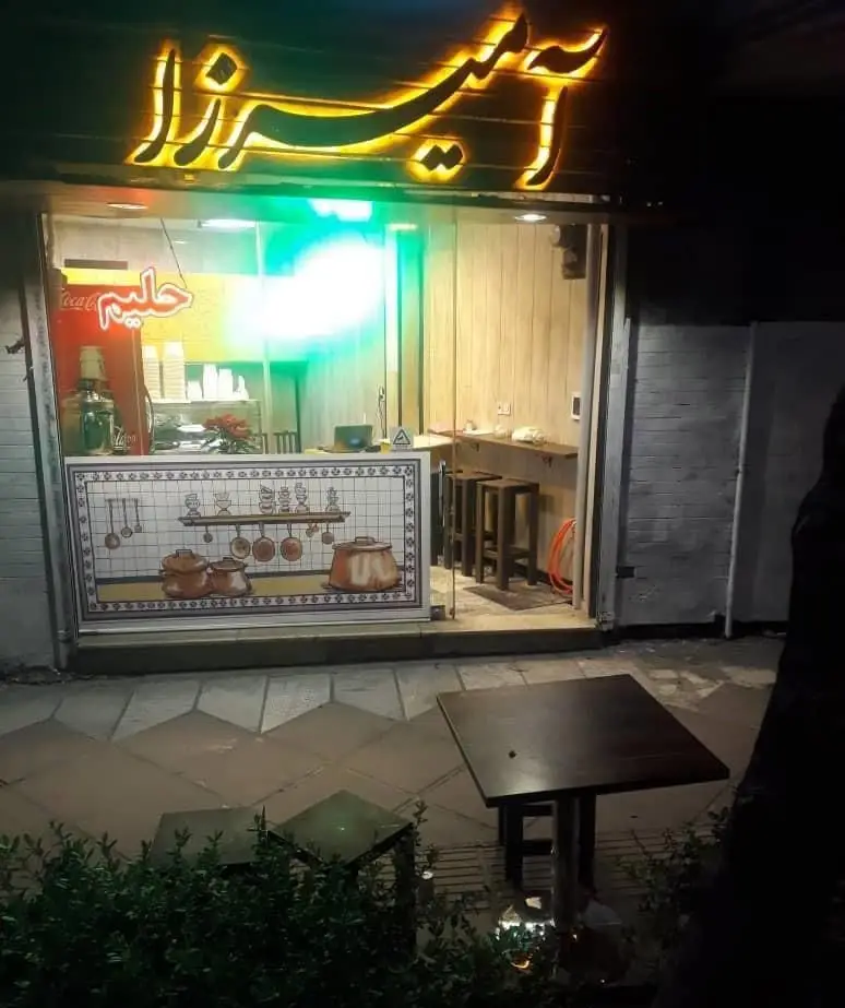 نمای شبانه آش و حلیم پزی آمیرزا با تابلوی نئون روشن، یخچال حلیم، دکوراسیون داخلی سنتی و میزهای فضای باز برای مشتریان.