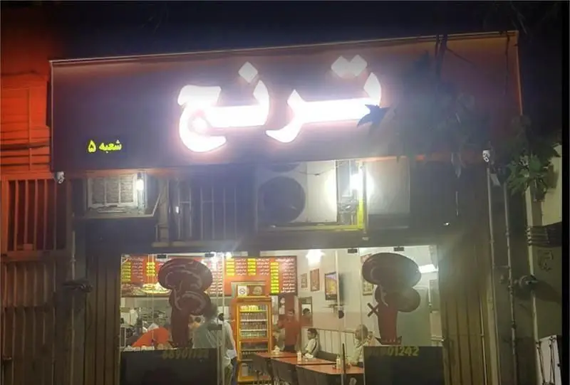 نمایی شبانه از پیتزایی ترنج شعبه ۵ با تابلوی روشن، دکوراسیون داخلی شامل مشتریان و منوی قیمت در ویترین شیشه‌ای فروشگاه.