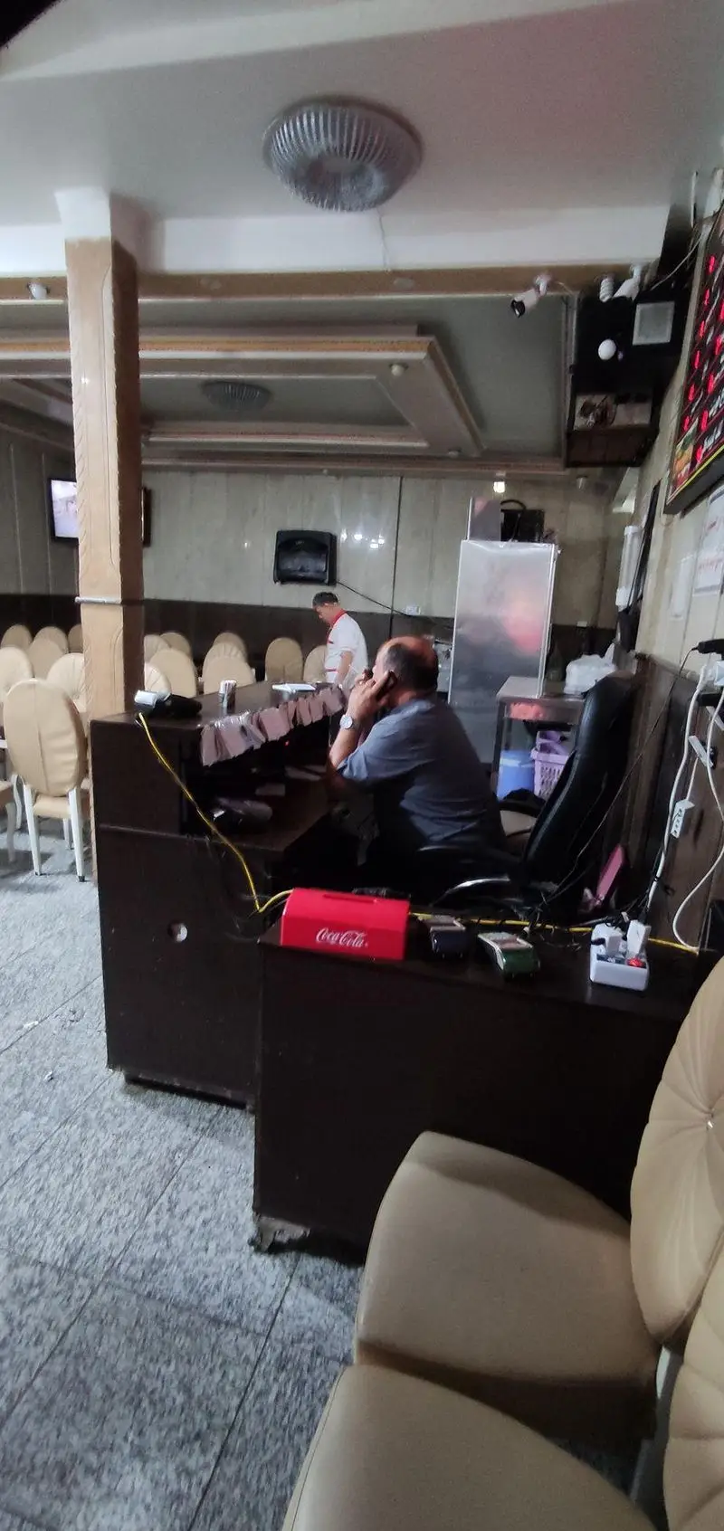 تصویری از داخل رستوران ایران زمین، شامل کانتر پذیرش، مشتری مشغول گفتگو با تلفن، و صندلی‌های خالی آماده پذیرایی از مهمانان.