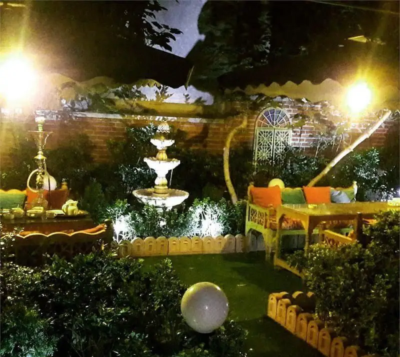 تصویری زیبا از باغچه شبانه کافه سفره خانه فارا با آبنمای سه‌طبقه، قلیان و چای سنتی، نورپردازی دلنشین و مبلمان راحت.