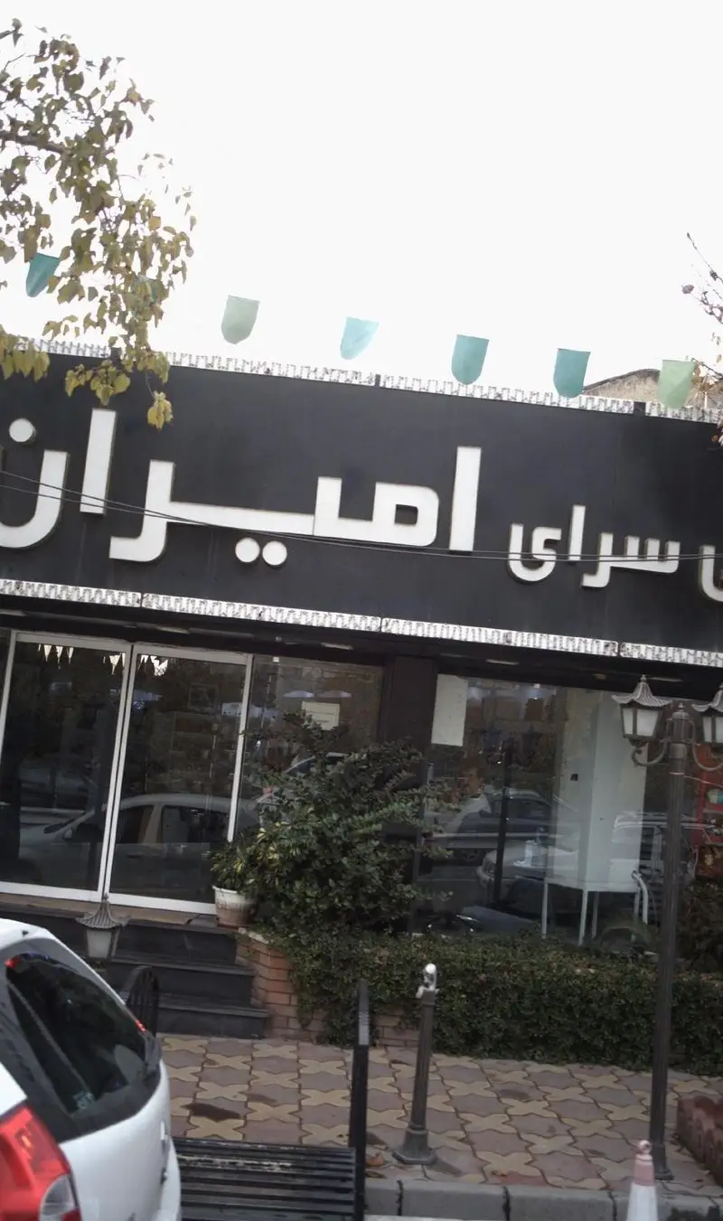 تصویر نمای بیرونی فروشگاه شیرینی سرای امیران، تابلوی بزرگ مشکی با نام سفید، ورودی شیشه‌ای، پیاده‌رو و فضای سبز اطراف.