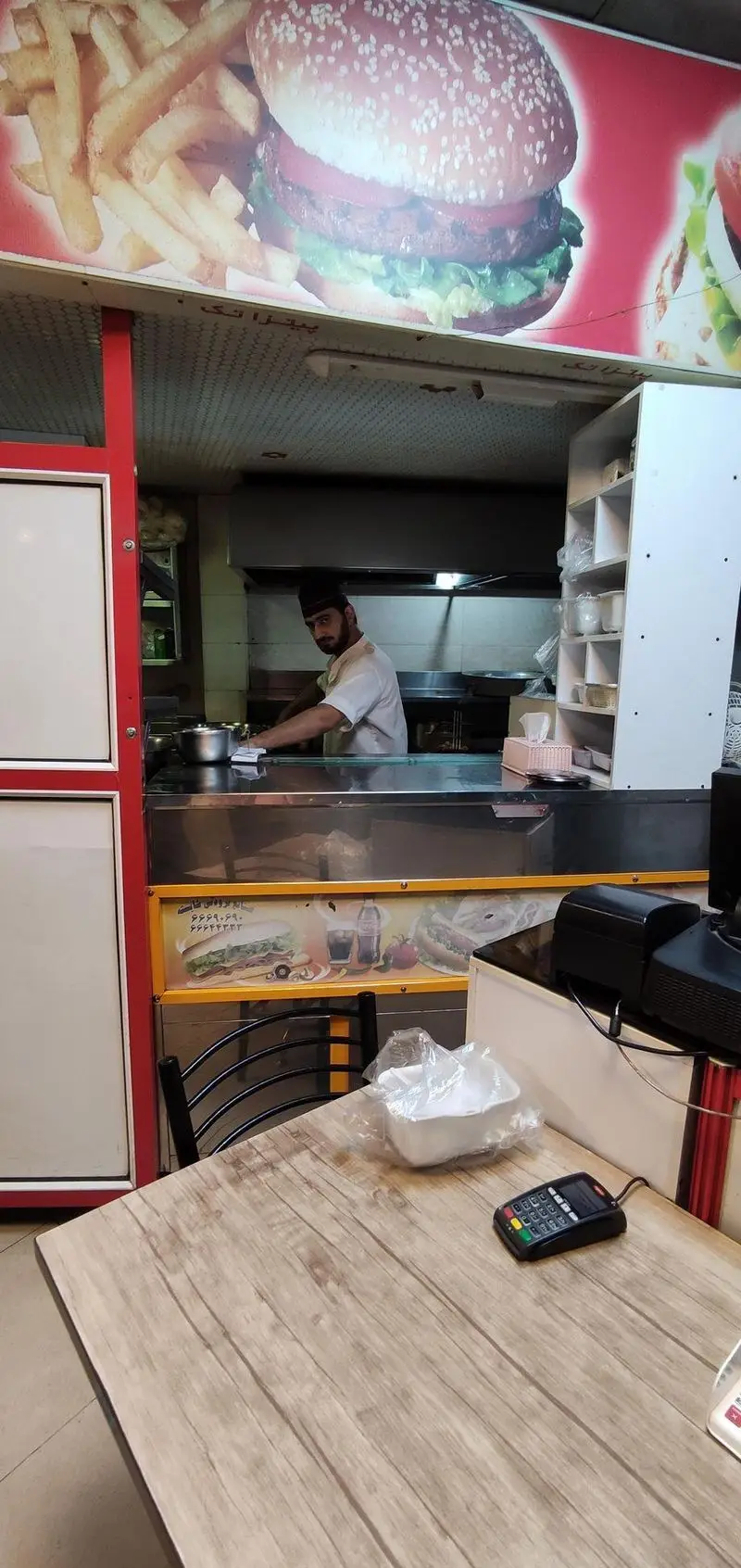 نمای داخلی فست فود پیتزا تک: آشپز مشغول کار در آشپزخانه، بنر بزرگ برگر و سیب زمینی و میز مشتری با دستگاه کارتخوان.