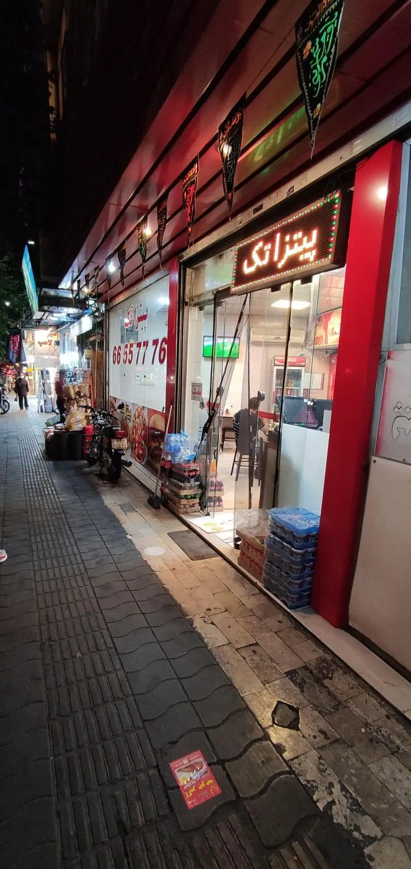 نمای شبانه رستوران فست فود "پیتزا تک" با تابلوی نئون درخشان، شماره تماس 66557776، و پرچم‌های مذهبی، جنب پیاده‌رو شلوغ.