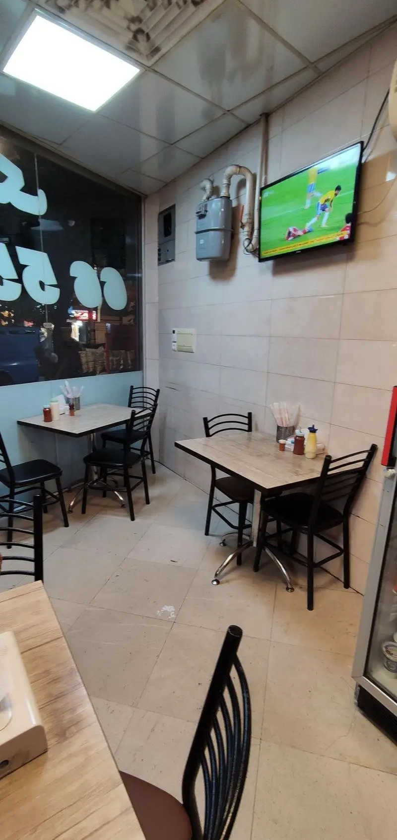 تصویری از فضای داخلی تمیز و دنج پیتزا تک با میز و صندلی‌های مرتب و تلویزیون در حال پخش فوتبال، محیطی ایده‌آل برای صرف غذا.