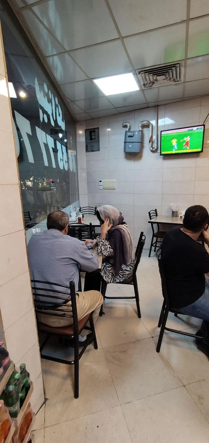 نمایی از فضای داخلی پیتزا تک با نورپردازی مناسب، میز و صندلی‌ها و مشتریان مشغول تماشای تلویزیون، با انعکاس نام پیتزا تک در شیشه.