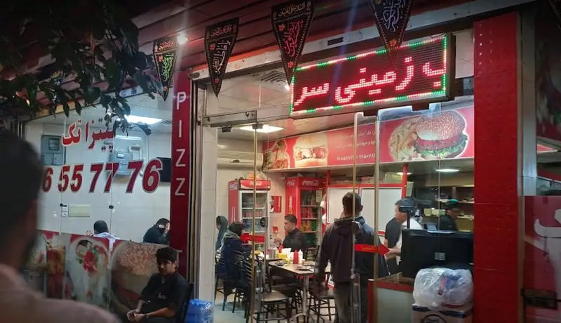 نمای بیرونی رستوران فست فود پیتزا تک با تابلوی روشن، شماره تماس ۶۵۵۷۷۷۶، و تبلیغ پیتزا، برگر و سیب زمینی سرخ کرده در شب.