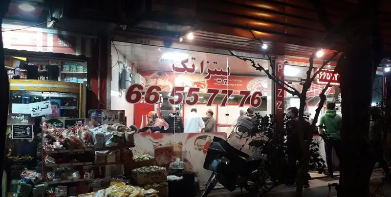 تصویر شبانه از پیتزا فروشی پیتزا تک با تابلوی روشن و شماره تماس 66557776، مکانی شلوغ برای سفارش انواع فست فود.