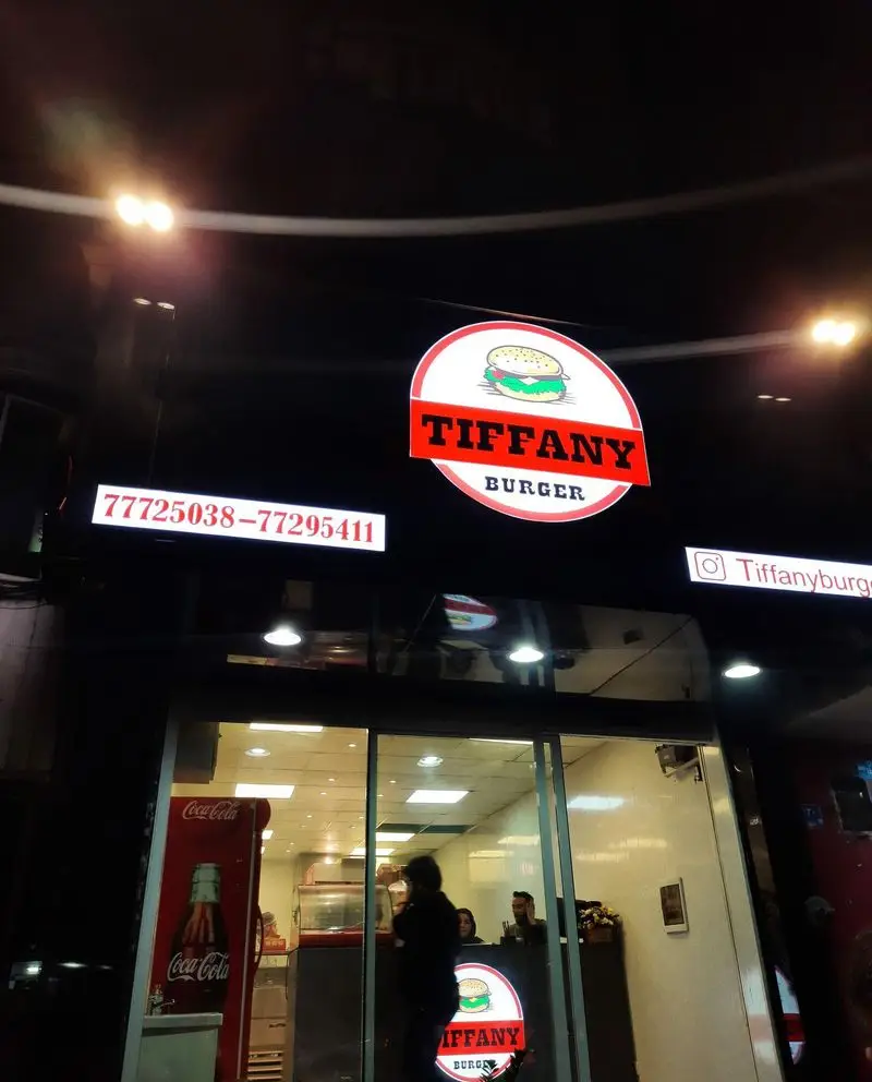 نمای شبانه فست فود تیفانی برگر با تابلوهای روشن شامل لوگو، شماره تماس 77725038-77295411 و آدرس اینستاگرام Tiffanyburger