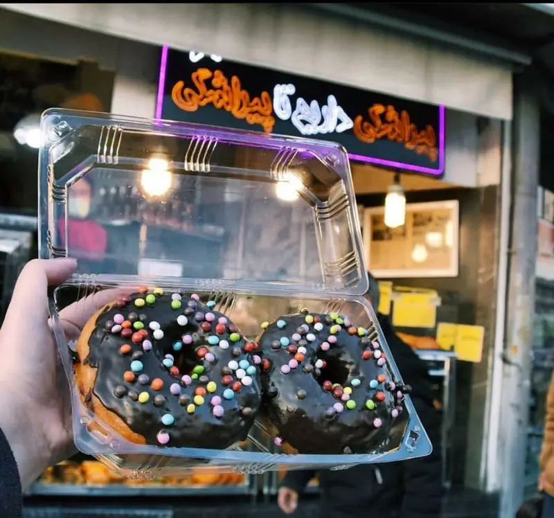 دو عدد دونات شکلاتی تازه با اسپرینکل‌های رنگی در جعبه شفاف، آماده برای لذت بردن از نان و پیراشکی بهمن تهران.