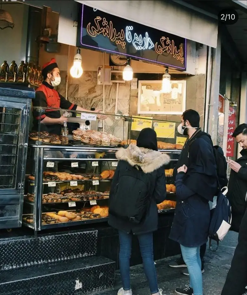 تصویری از مغازه نان و پیراشکی بهمن، ویترین پر از انواع پیراشکی، نان و شیرینی تازه با حضور فروشنده و مشتریان در خیابان.