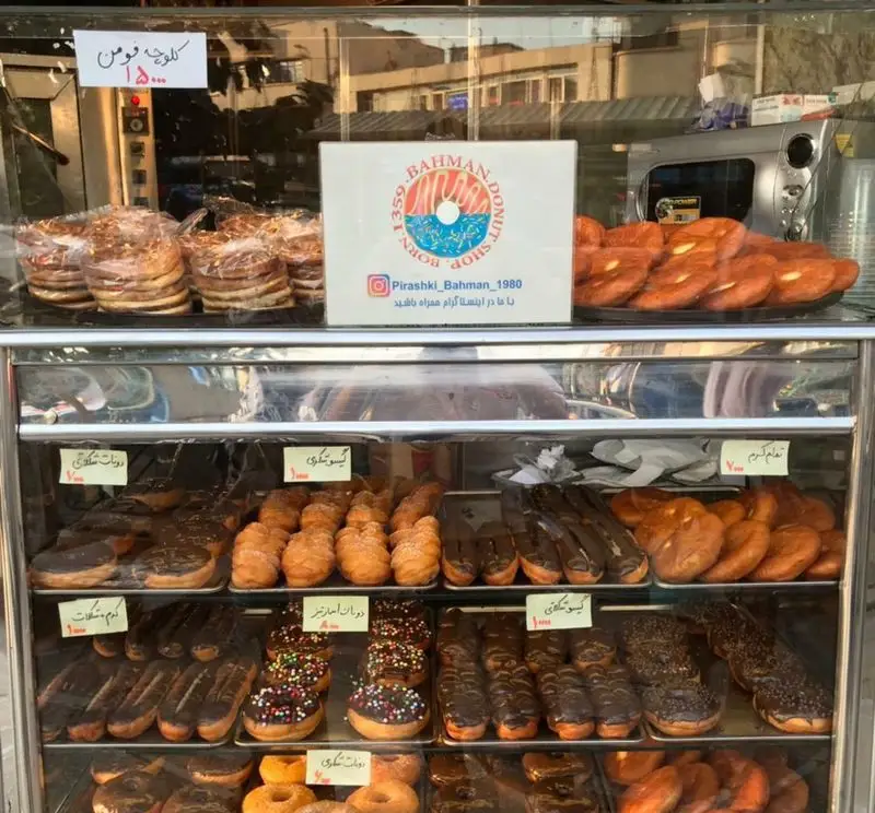 نمای نزدیک از ویترین نان و پیراشکی بهمن، پر از دونات‌های متنوع شکلاتی و رنگارنگ، کلوچه و شیرینی‌های تازه پخته.