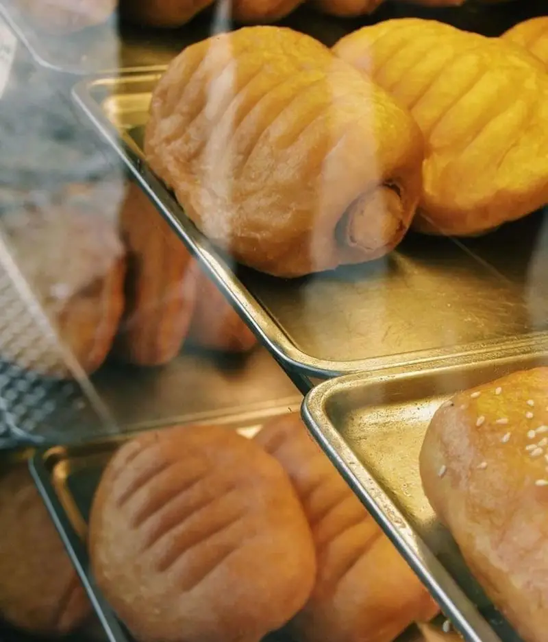 نان و پیراشکی‌های متنوع و تازه بهمن با رویه طلایی و شیاردار، چیده شده در سینی‌های فلزی براق پشت شیشه ویترین.