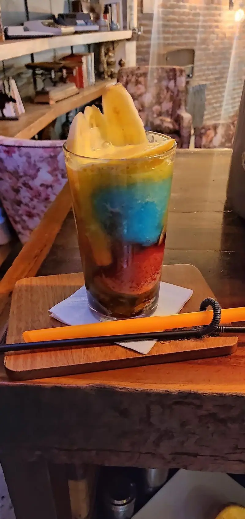 نوشیدنی خنک و رنگارنگ لایه‌ای با برش‌های میوه در لیوان بلند، روی سینی چوبی در کافه رستوران هوکر با محیطی دنج.