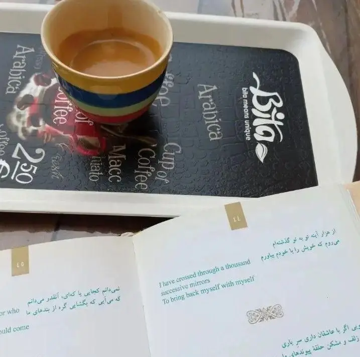 یک فنجان قهوه گرم در کافه روکو، کنار کتابی باز با اشعار فارسی و ترجمه انگلیسی، محیطی دلنشین برای مطالعه.