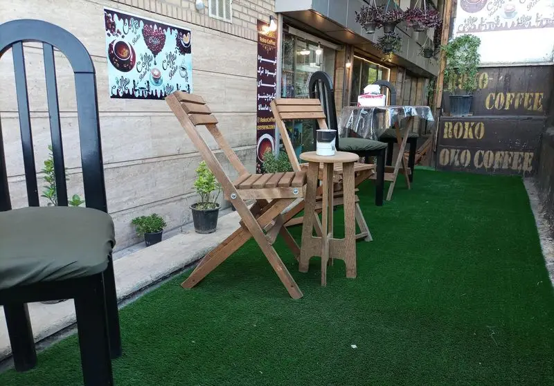 فضای باز کافه روکو با چمن مصنوعی سبز، میز و صندلی‌های چوبی شیک، محیطی دلپذیر برای لذت بردن از انواع قهوه و نوشیدنی.