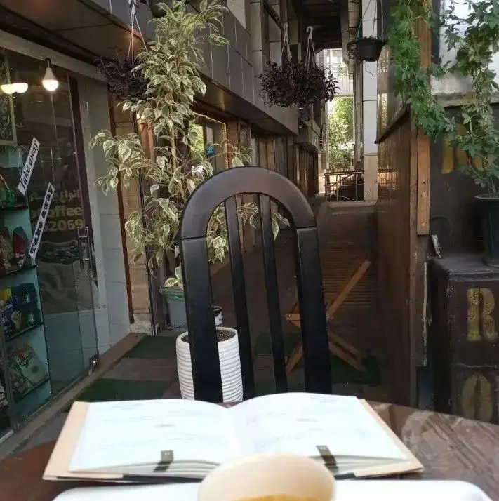 نمایی از فضای بیرونی کافه روکو با میز، صندلی مشکی، منوی قهوه، و گیاهان طبیعی. مکانی دنج برای لذت بردن از قهوه و خدمات بیرون‌بر.