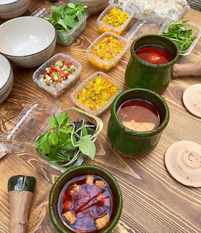 غذای سنتی دیزی رستوران دیزی دیدار با مخلفات متنوع شامل سبزی، سالاد شیرازی و نان تازه در ظروف سفالی روی میز چوبی.