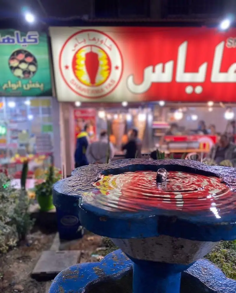 نمای بیرونی رستوران فست فود شاورما یاس در شب با تابلوهای روشن و لوگو شاورما، همراه با فواره‌ای زیبا در پیش زمینه.